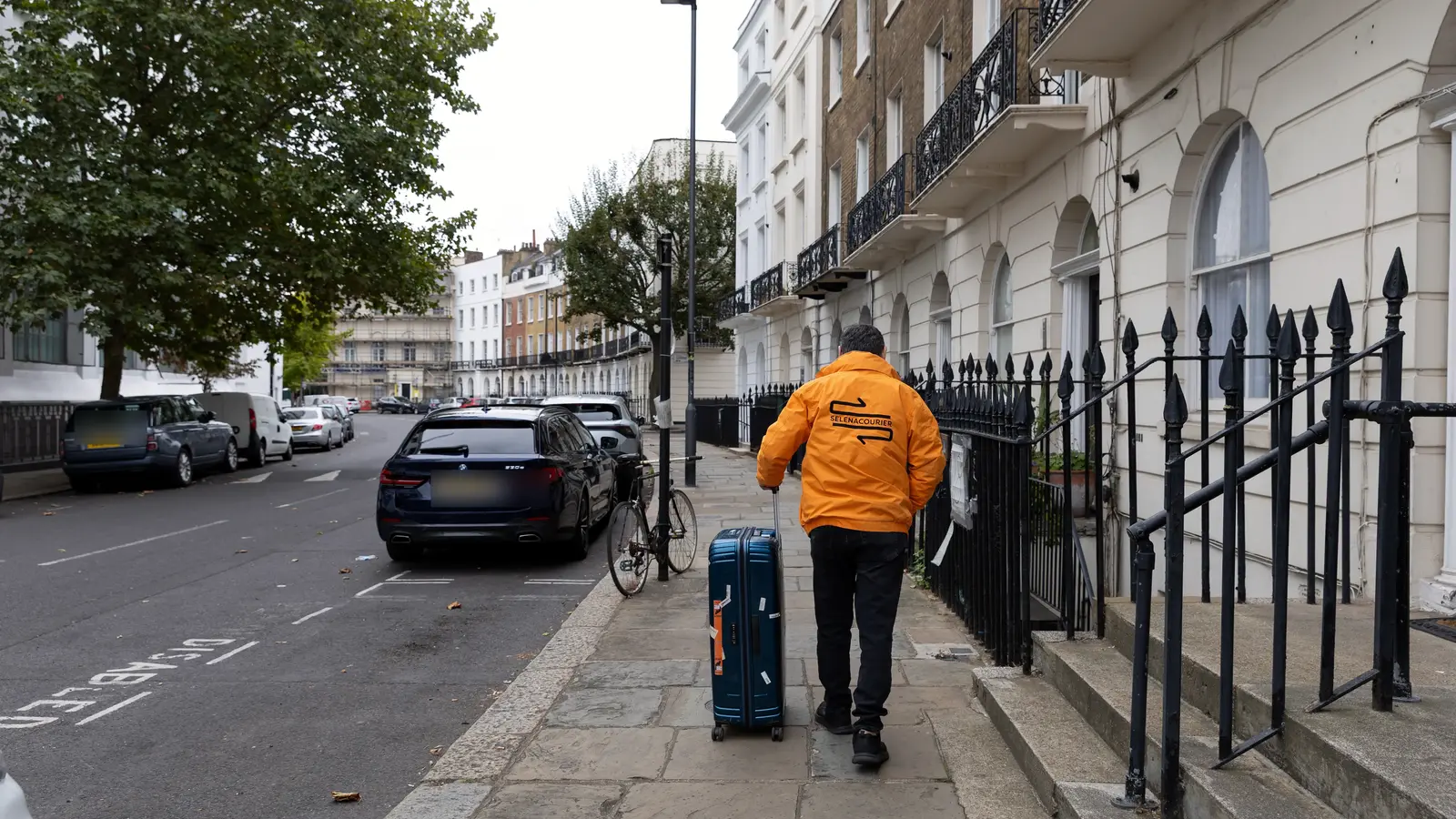 A Selena courier in an orange jacket wheels a navy suitcase along a London pavement lined with black railings